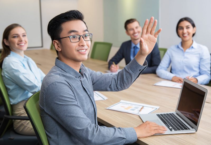 Young Asian businessman wearing glasses sitting at meeting or conference in boardroom with multiethnic colleagues and raising hand to ask question